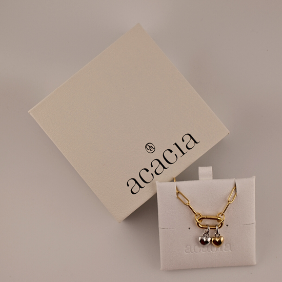 Gold necklace with heart pendant on a white card with 'acacia' branding, against a beige background.