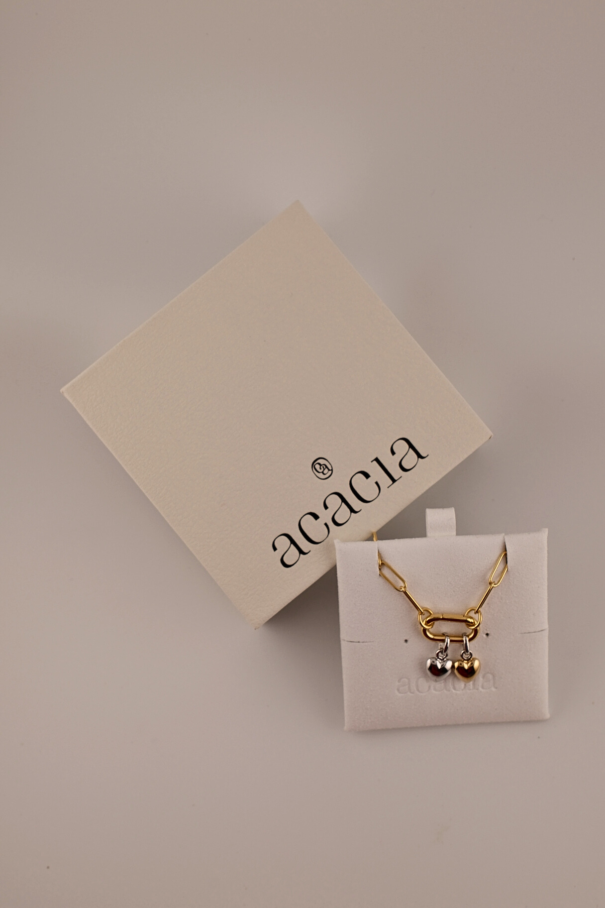Gold necklace with heart pendant on a white card with 'acacia' branding, against a beige background.